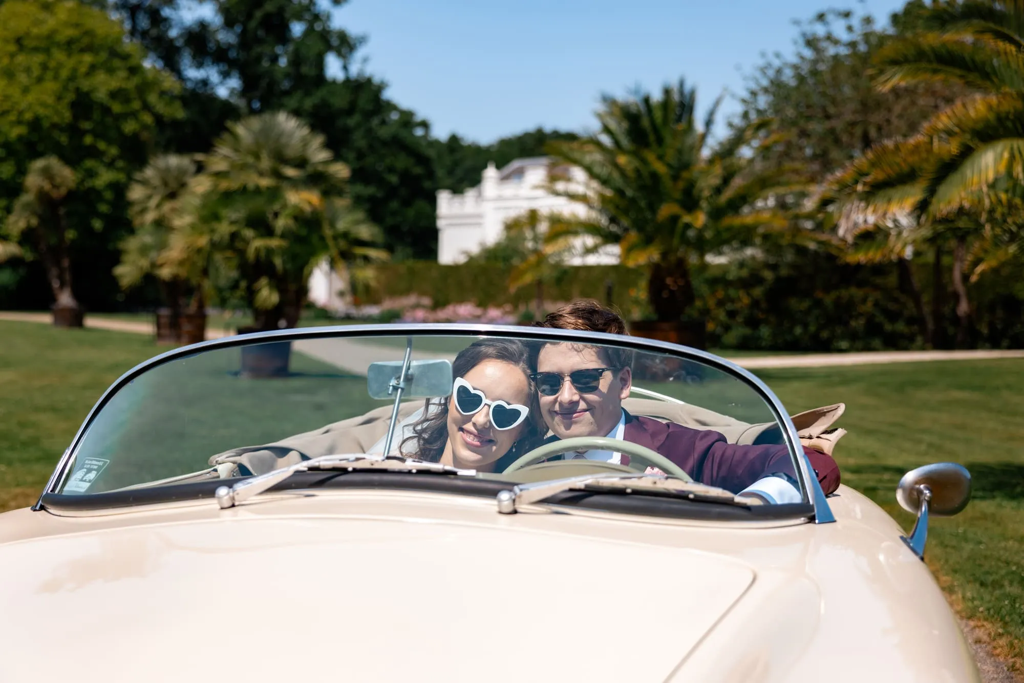 Het bruidspaar rijdt in een crèmekleurige cabriolet, allebei met een hartjeszonnebril op