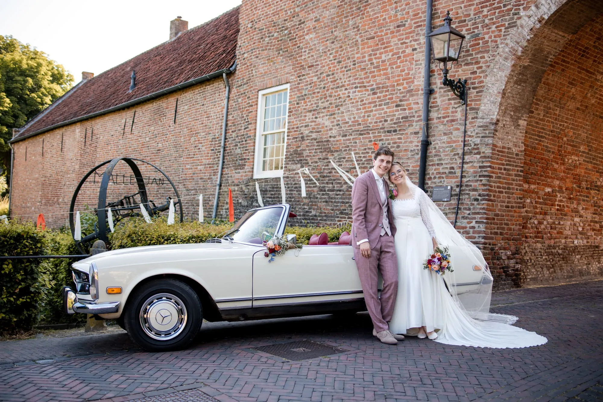 De bruid staat bij een klassieke witte Mercedes voor een oude kloostermuur
